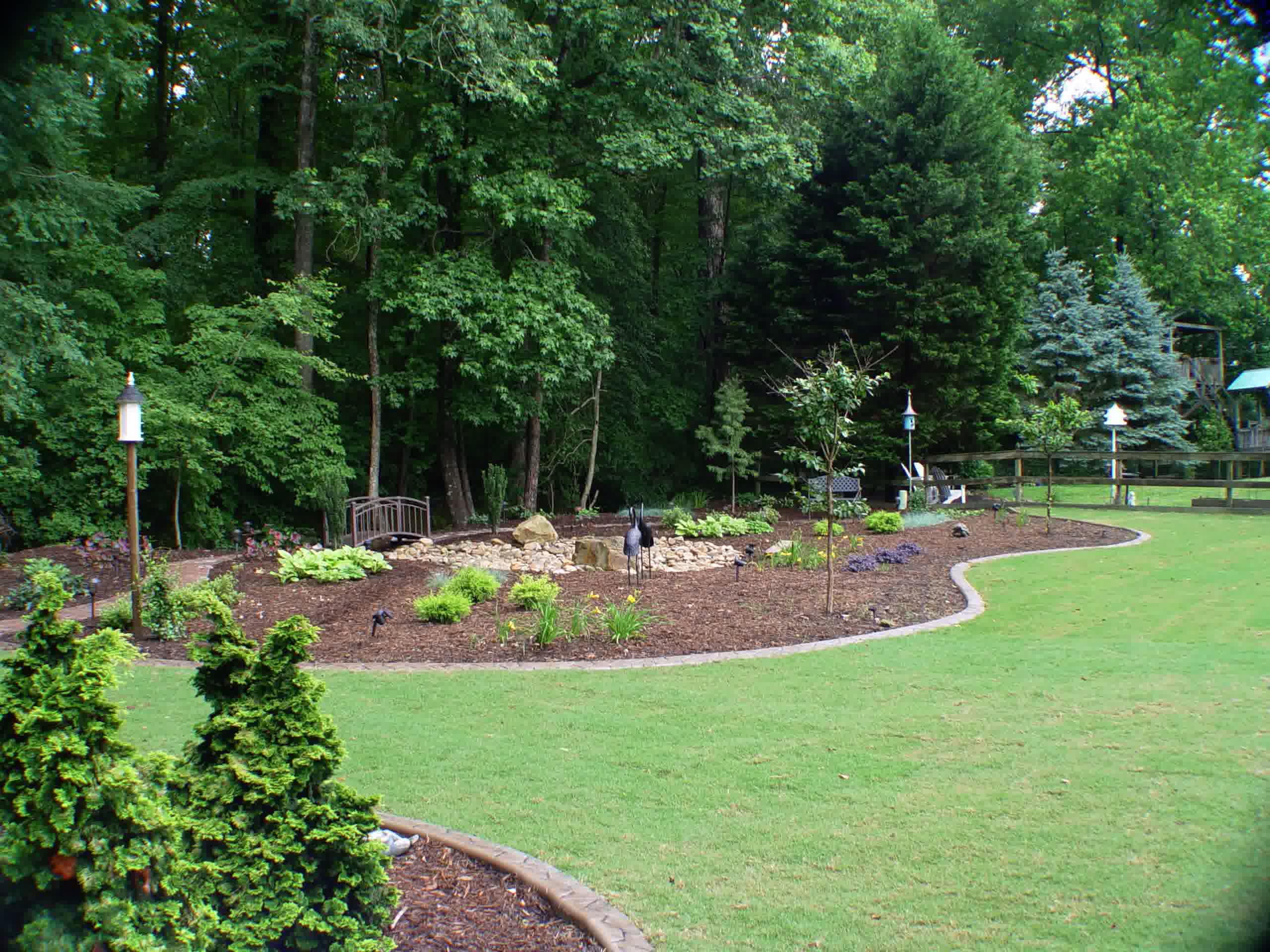 solomons yard cleanups reidville sc - Solomon's Landscape & Design Cleaned and mulched backyard garden with trimmed shrubs, landscape lighting, and stone border along a forest edge.